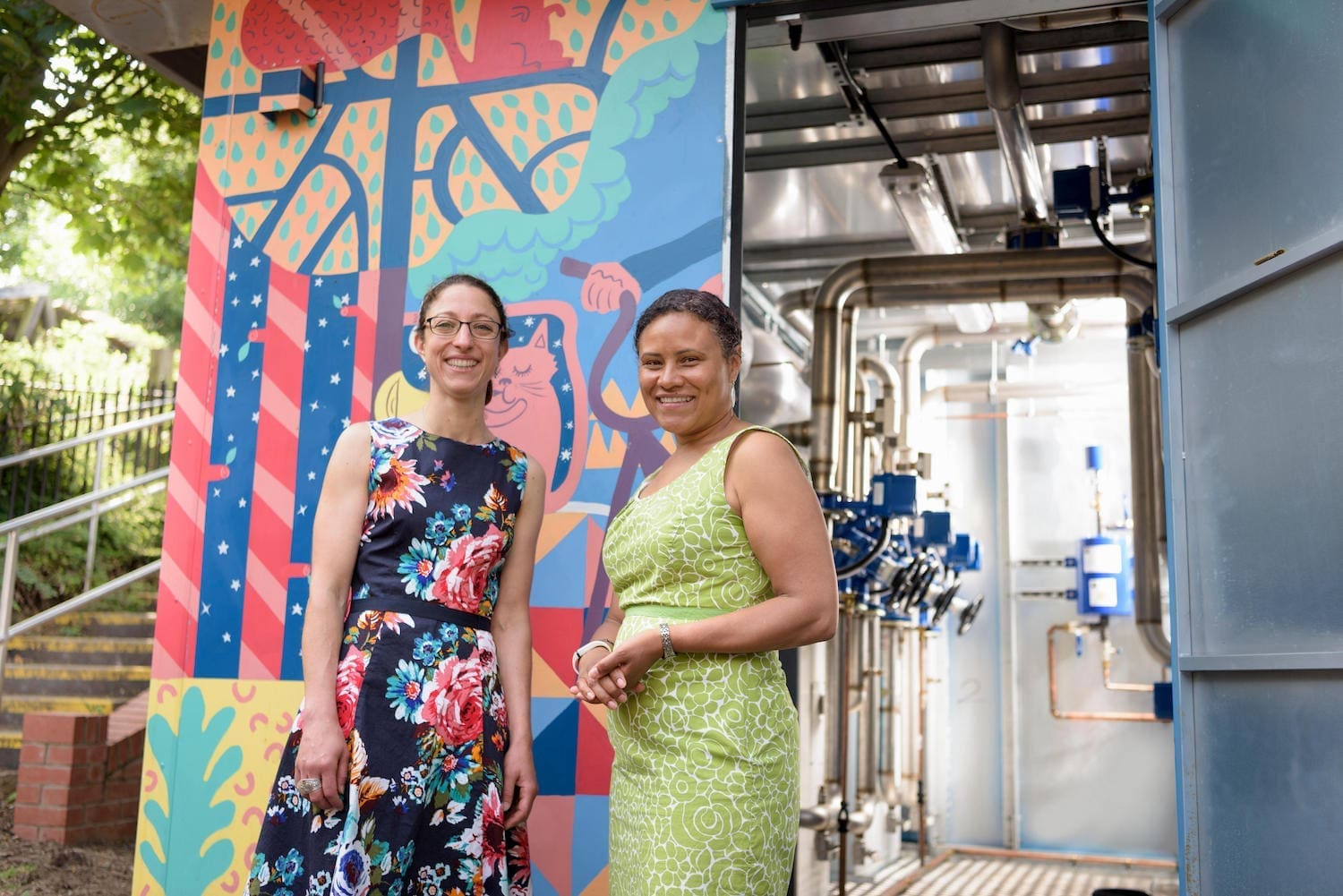 Two women standing in front of a pumping station in the middle of an urban setting. The outside of the pumping station is painted with cool artwork. One  woman is wearing a blue  floral dress and glasses, the other woman is wearing a green floral dress.